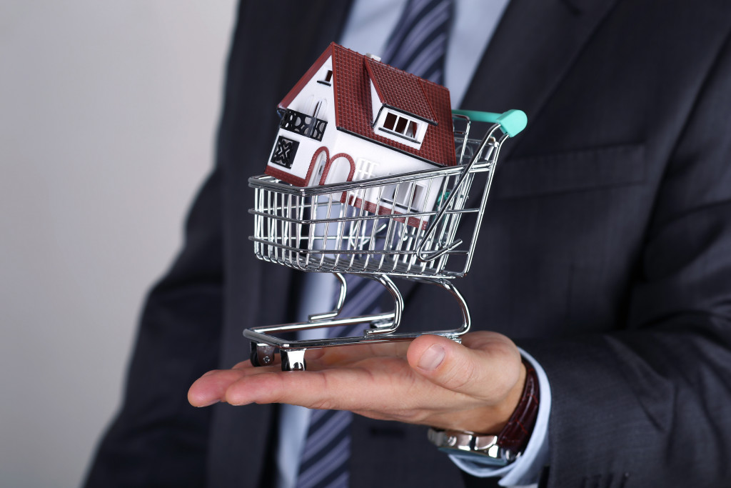 a male property seller holding a miniature grocery cart with a house in it